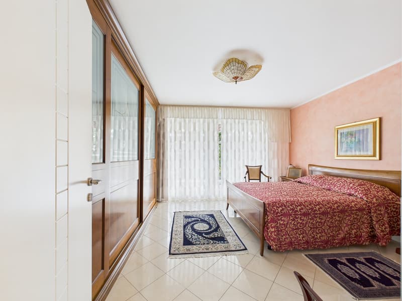 Bedroom with a double bed, wooden headboard, pink walls, white ceiling, white tiled floor, two rugs, a chair, a nightstand, a picture frame, a lamp, and a window with white curtains.