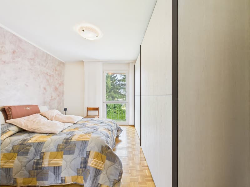 Bedroom with bed, pillows, bedside lamp, chair, glass door, balcony, wooden floor, white walls, ceiling lamp