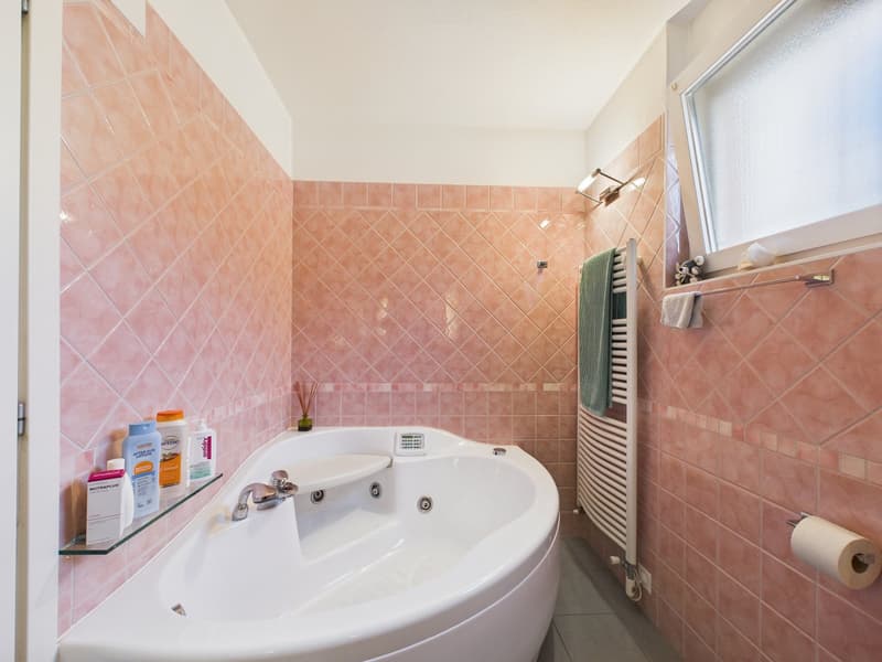 Bathroom with pink tiles, large bathtub, glass shelf, towel rack, window, and toiletries