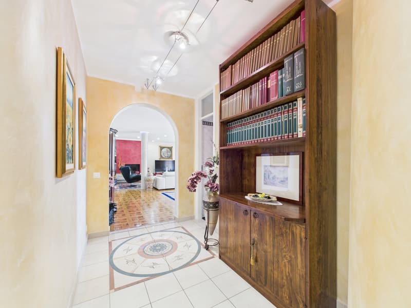 A hallway with a wooden bookshelf, a round carpet, and a vase with flowers. The hallway leads to a living room with a couch, a television, and a fireplace.