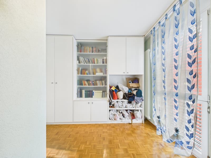 Empty room with wooden floor, white walls, white cabinets, glass door cabinet with books, white curtains, and a radiator