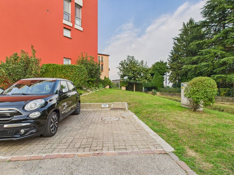 paved parking area, black car parked, red building, green grass, bushes, trees