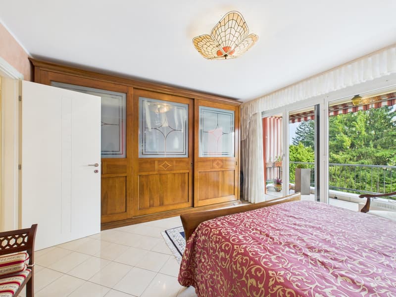 Bedroom with wooden wardrobe, white door, balcony, bed with pink bedspread, wooden chair, tiled floor, decorative lamp