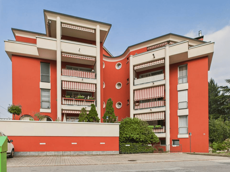 3 story building, red exterior, white trim, balconies, windows, plants, trees, parking area