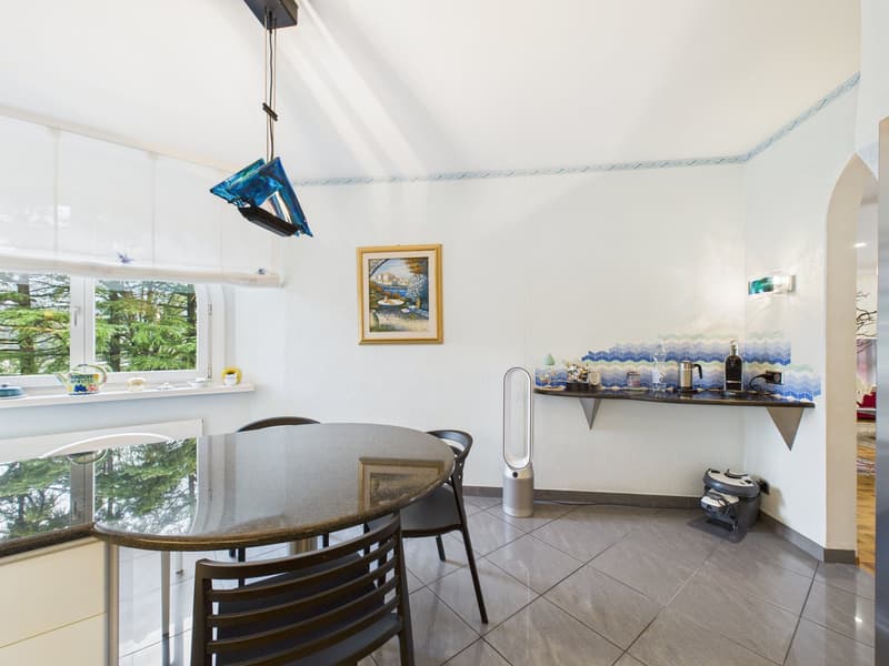 Dining area with a round glass table, black chairs, a picture frame on the wall, a window with a view of trees, a shelf with a kettle, a bottle, and a lamp, and a white wall with a blue border.