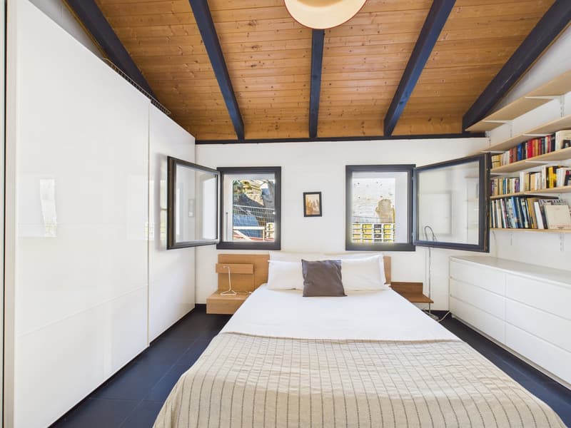modern bedroom with wooden ceiling, white walls, black floor, bed with pillows, bedside table, open windows, wooden bench, white cabinets, shelves with books