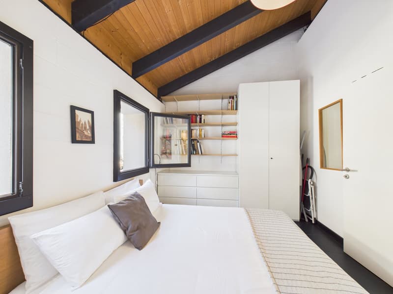 Bedroom with a bed, wooden ceiling, white walls, white bedspread, white pillows, a shelf with books, a white cabinet, a window, and a picture frame.