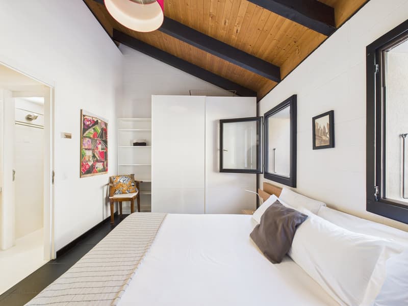 Bedroom with a bed, wooden ceiling, white walls, large windows, a chair, a shelf, and a lamp