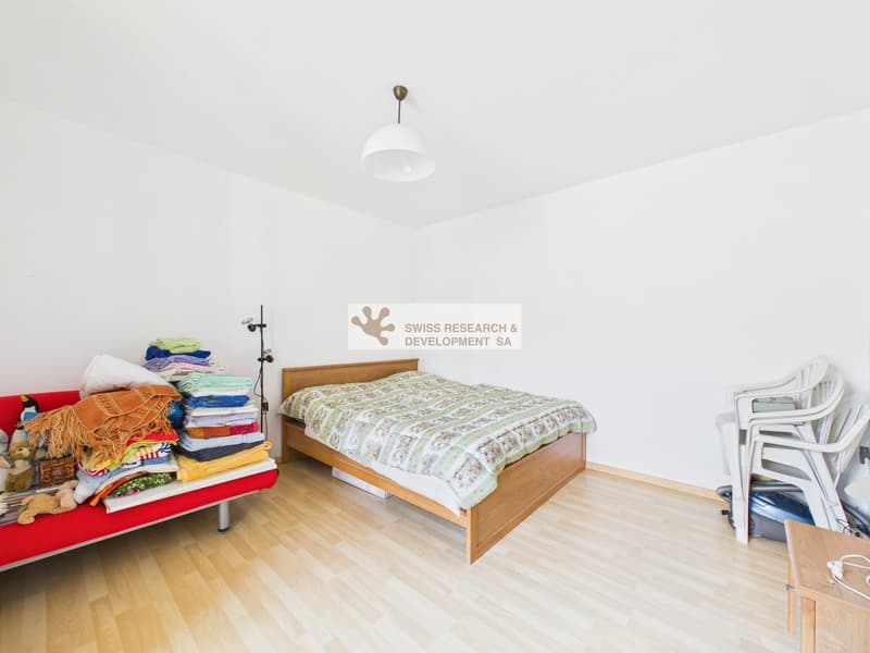 A room with a bed, a couch, a lamp, and a stack of clothes on the couch.