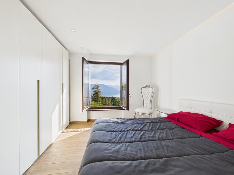 modern bedroom, white walls, wooden floor, large window with balcony, bed with red pillows, white chair, white wardrobes