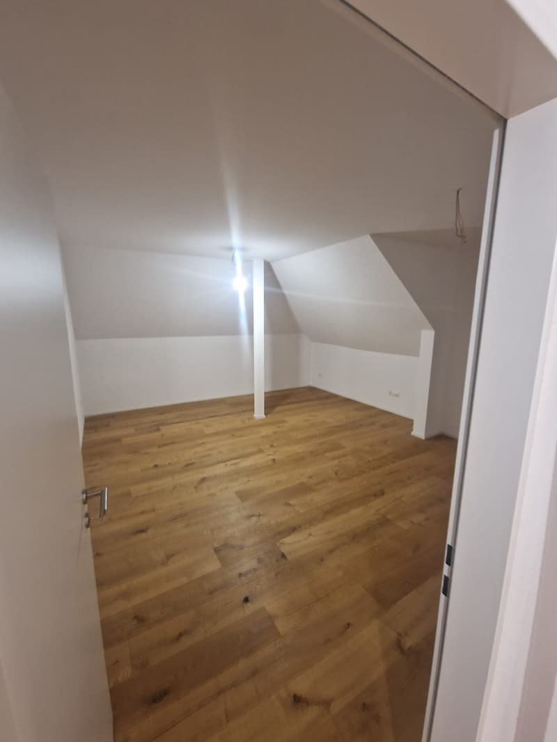 Empty room, wooden floor, white walls, slanted ceiling, light reflecting off the floor, power outlet