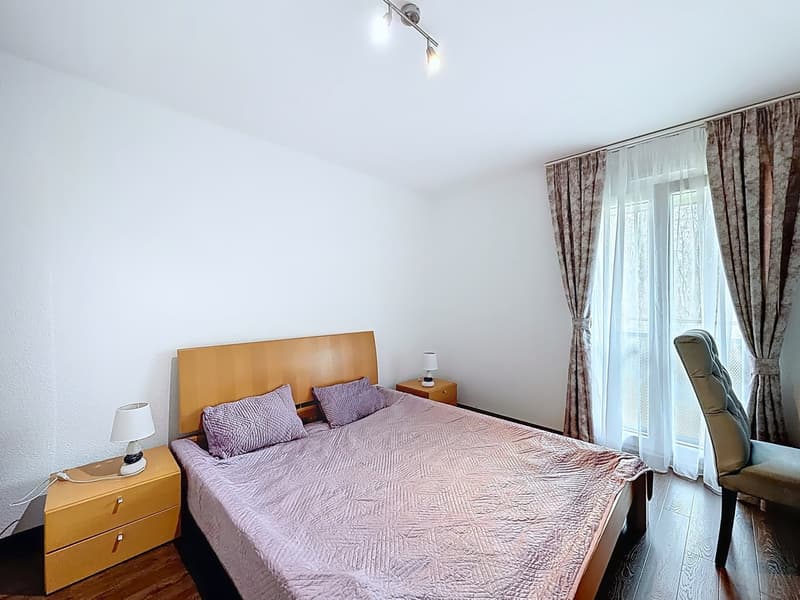 A bedroom with a bed, two nightstands, lamps, and a chair. The room has a window with curtains.