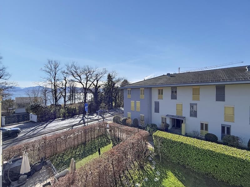 A two-story house with yellow shutters, a gray roof, and a garden in front. The house is surrounded by trees and bushes, and there is a car parked on the side of the road. In the distance, there is a mountain range and a body of water.