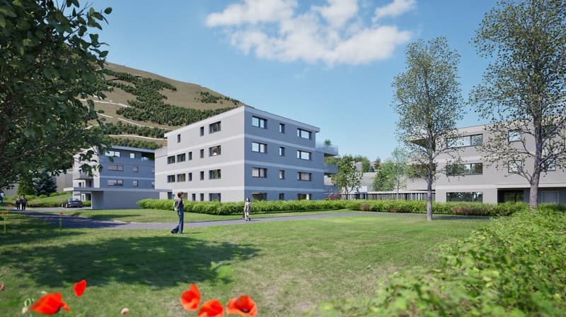 A modern, multi-story apartment building with a gray exterior and large windows. The building is surrounded by a grassy area with trees and flowers in the foreground. There appear to be balconies or terraces on some of the units.