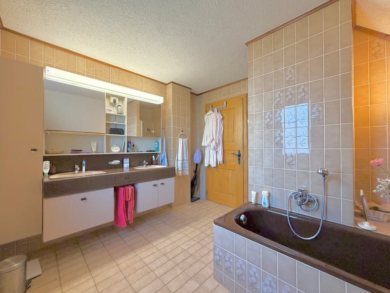 Two sinks, bathtub, mirror, wooden door, white cabinets, tile flooring, light fixtures