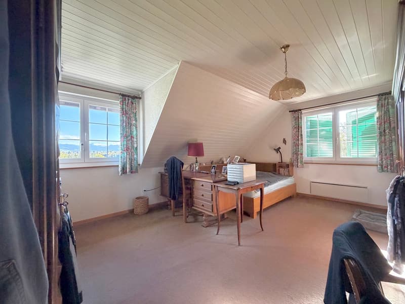 Carpeted floor, wooden desk, lamp, window, curtain, bed, radiator, wicker basket, framed photos, stack of books, and lamp