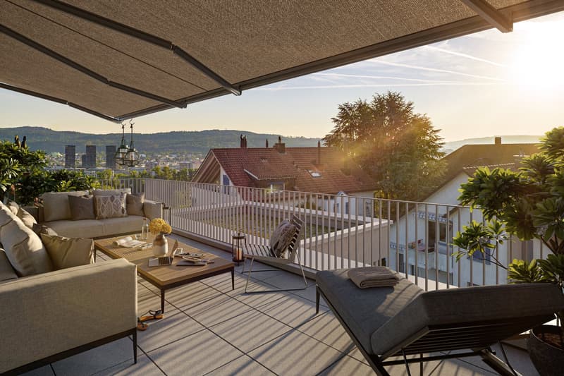 Spacious balcony with comfortable outdoor furniture, including a sofa and chairs, overlooking a scenic cityscape with mountains in the background. The balcony is covered by a retractable awning and features potted plants and greenery, creating a relaxing