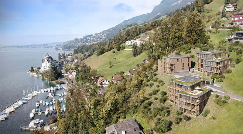 This image shows a scenic view of a lakeside town nestled in the mountains. The town features a mix of traditional and modern architecture, with a historic castle-like building and newer apartment buildings with green roofs. The town is surrounded by lush