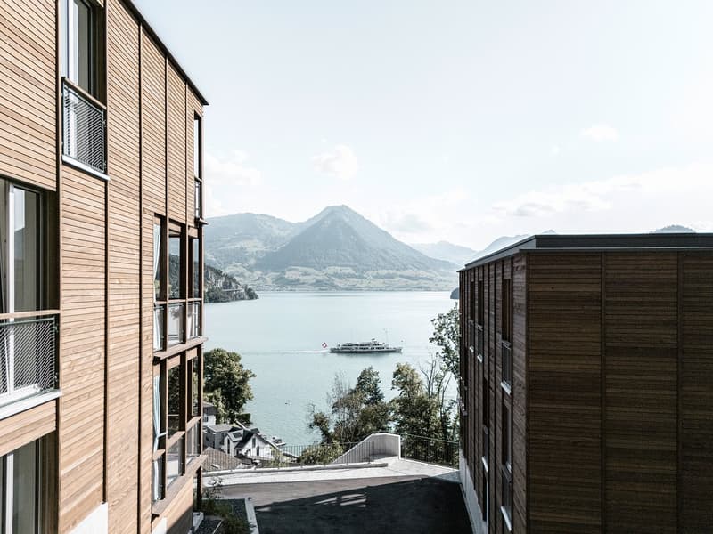 The image shows a multi-story wooden building with large windows overlooking a scenic lake and mountains in the background. A boat can be seen sailing on the lake. The building appears to be situated on a hillside, with a paved path or road leading down t