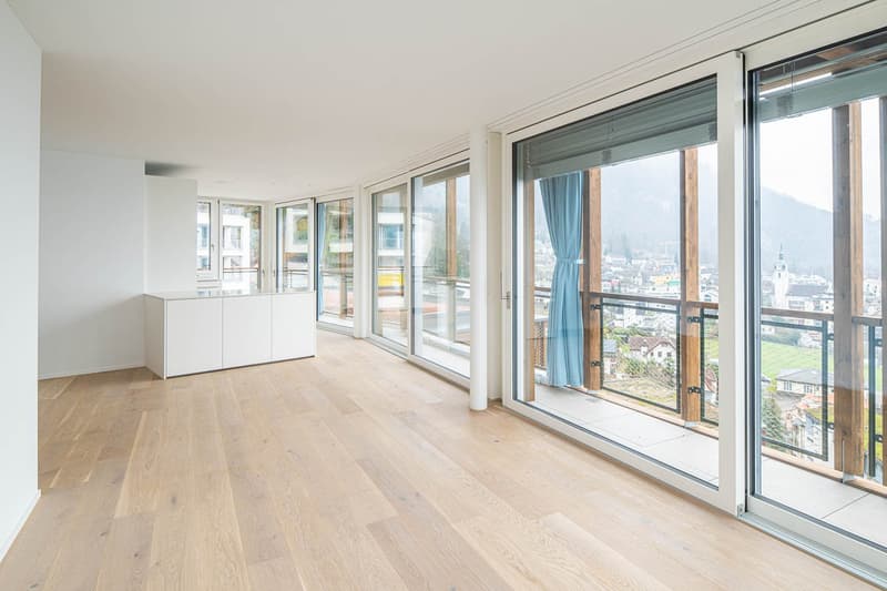 Spacious living room with large windows and sliding glass doors leading to a balcony, offering a scenic view of the city. The room has a minimalist design with light-colored hardwood floors and white walls. There is a built-in cabinet unit along one wall.