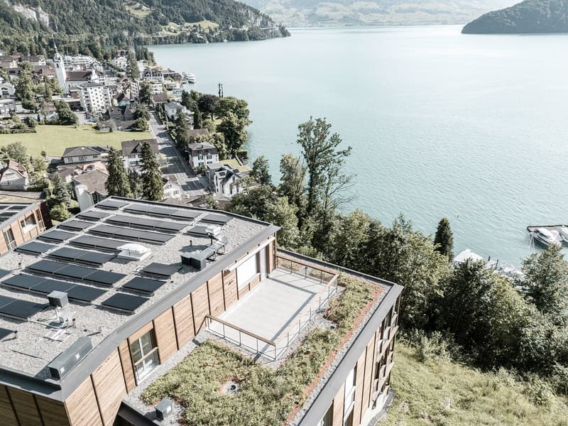 Multi-story building with a green roof and balconies overlooking a scenic lake surrounded by mountains and forests