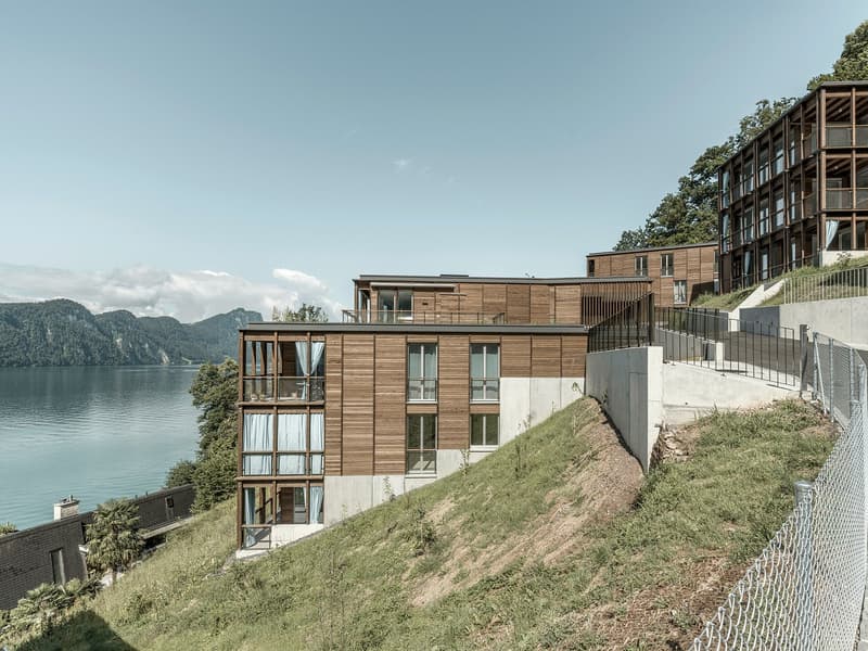 Multi-story apartment building with wooden exterior, large windows, and balconies overlooking a lake and mountains in the background. The building is situated on a hillside with a grassy slope leading down to the water's edge.