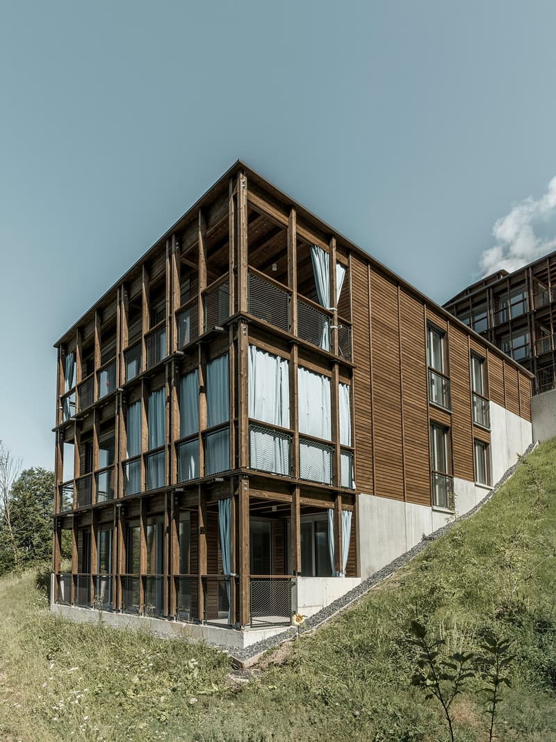 A modern, multi-story building with a wooden frame and large windows. The building has a balcony or terrace on one side.