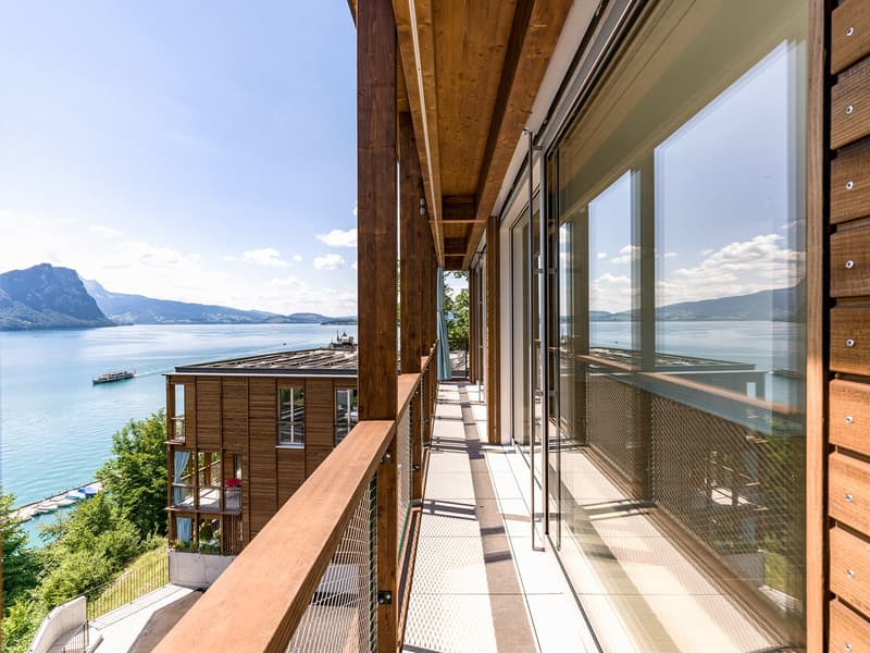 Multi-story wooden building with large windows and a balcony overlooking a lake and mountains in the background. The building has a wooden deck and railing system along the exterior.