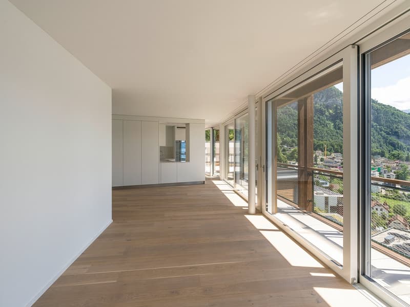 Spacious open-plan living area with large windows offering a scenic view of the surrounding mountains and landscape. The room features hardwood flooring, neutral-colored walls, and built-in cabinetry along one wall.