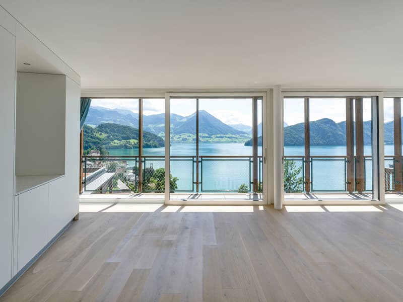 Spacious living room with large windows offering a panoramic view of a lake surrounded by mountains. The room has a minimalist design with light-colored hardwood floors and white walls. The windows provide abundant natural light and a serene, picturesque