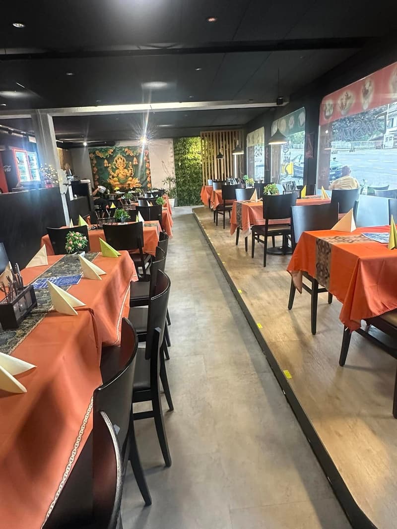 Restaurant with tables and chairs, orange tablecloths, black chairs, wooden floor, plants, and large windows