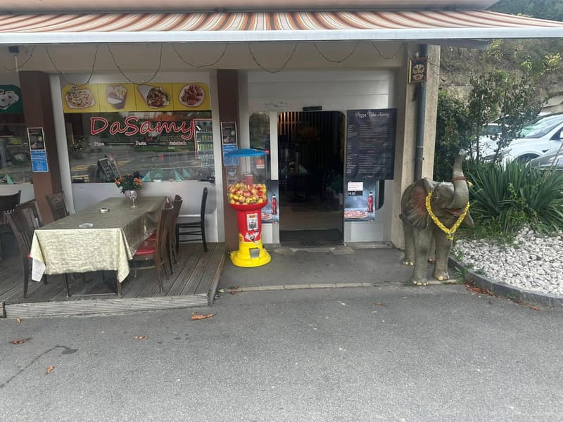 A small storefront with a wooden deck, tables and chairs for outdoor seating, a candy machine, and a small decorative elephant statue.