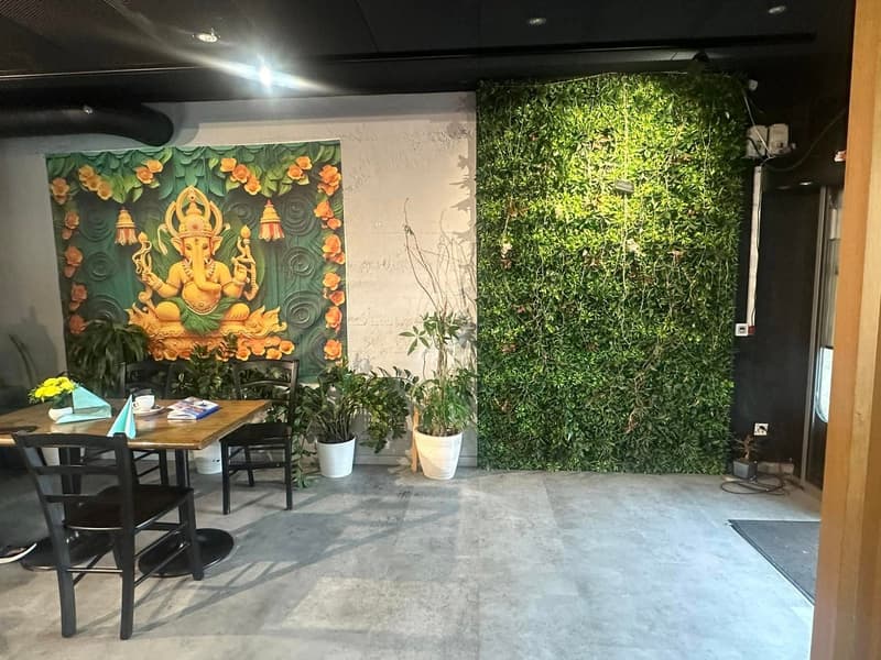 a restaurant area with a green plant wall, a decorative wall mural, wooden dining table, black chairs, white ceramic pots, yellow flowers
