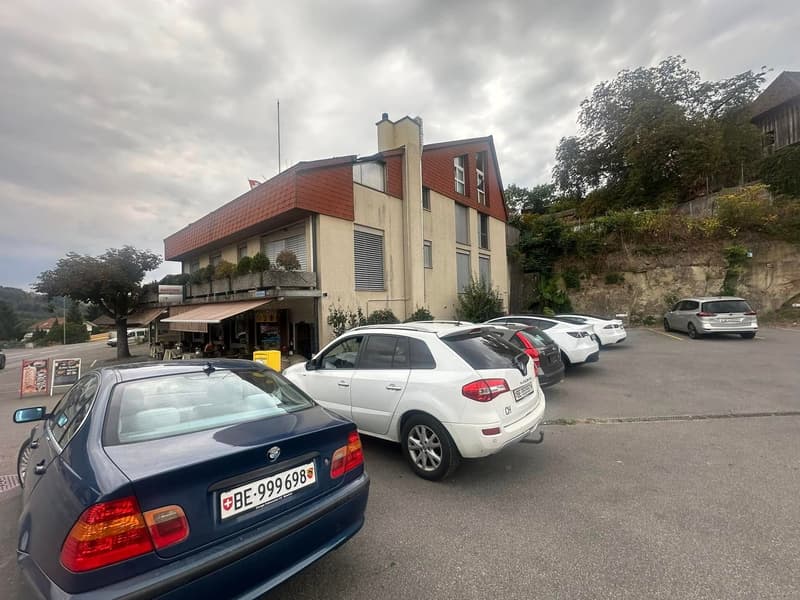 Parking area in front of a building, cars parked, building has awnings, flag on top