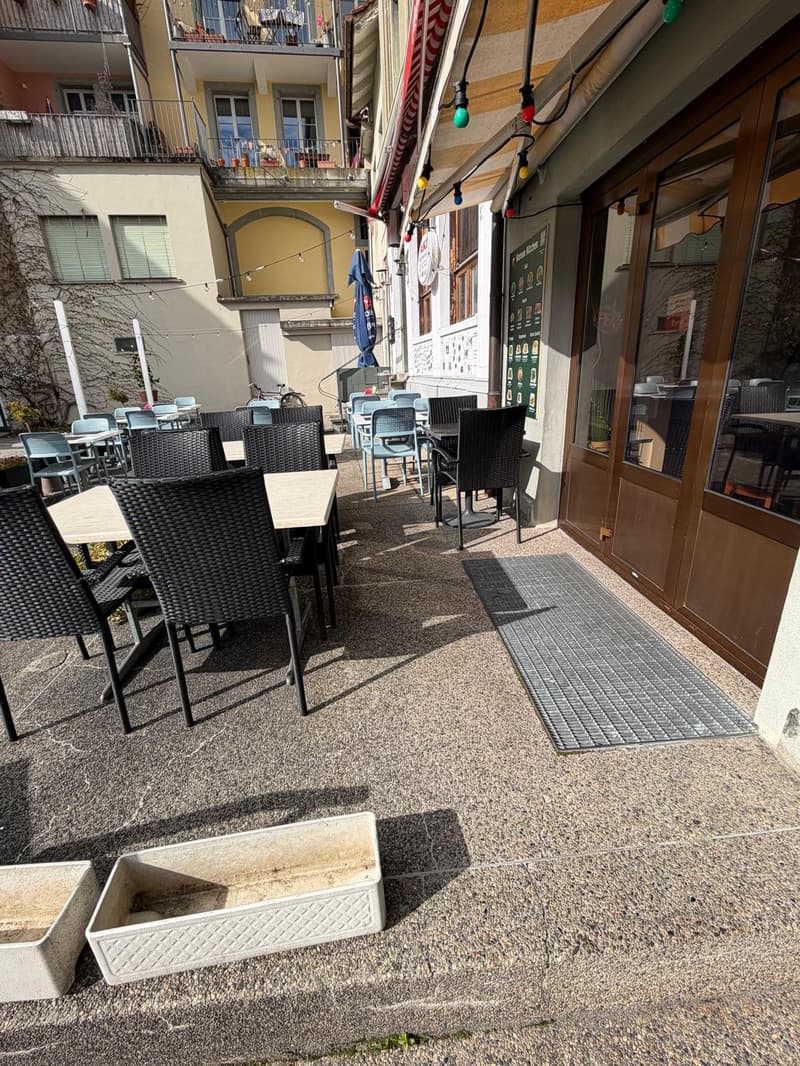 Outdoor seating area with tables and chairs, concrete floor, metal grate, potted plants, awning, and decorative lights.