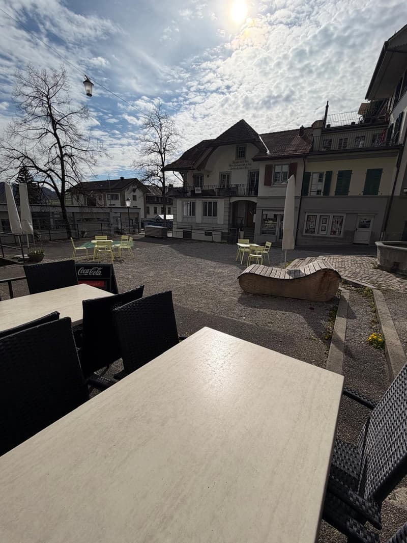 Outdoor seating area with tables and chairs, an open space with a building in the background, and a bench