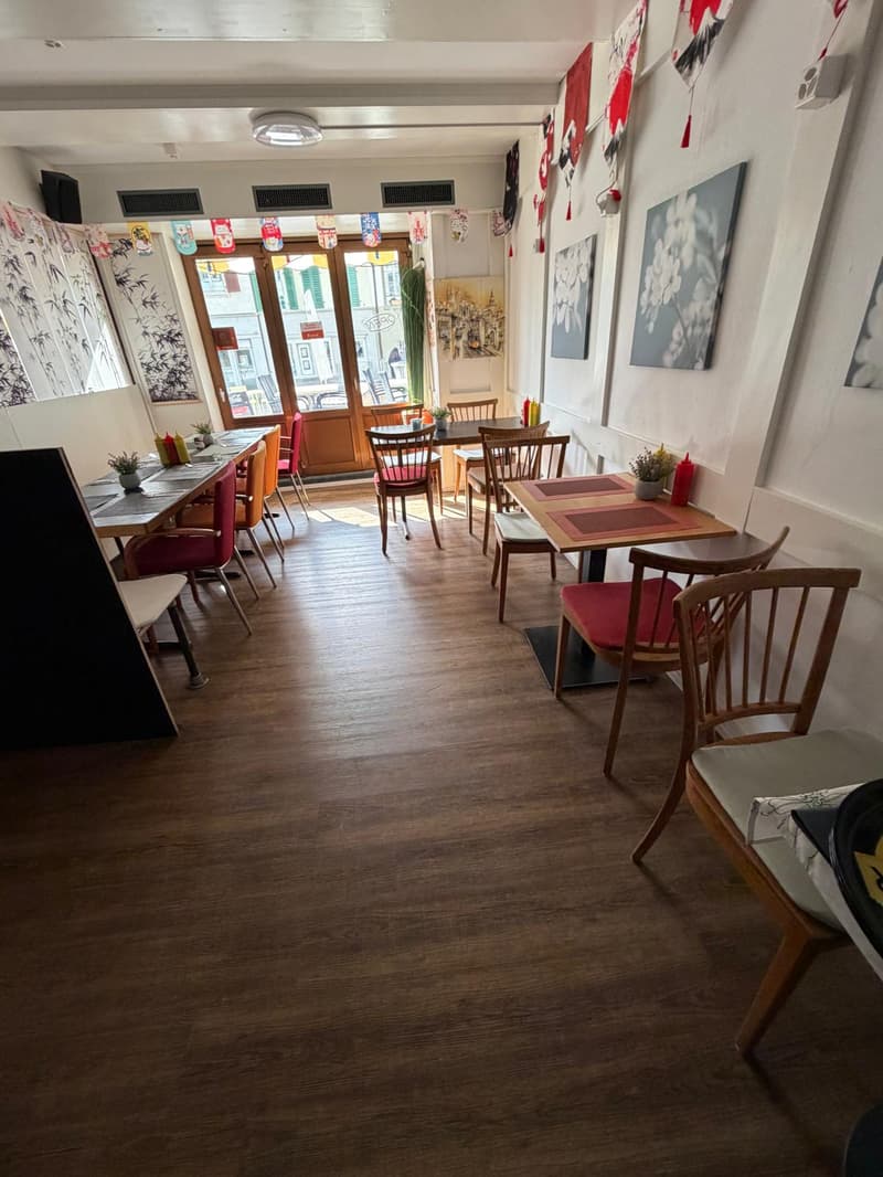 empty restaurant with wooden floor, wooden tables and chairs, white walls, decorative items, and a glass door leading to the outside