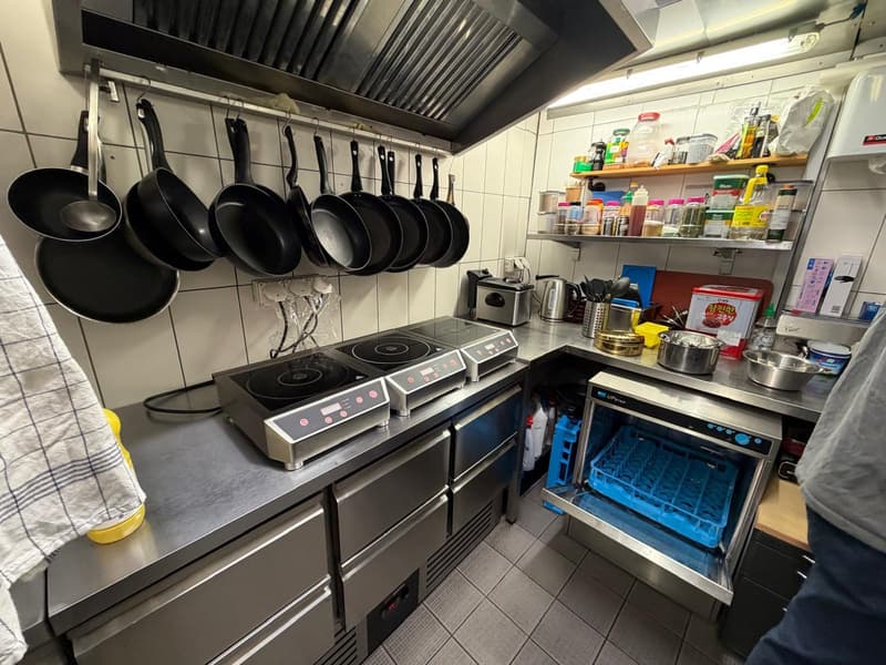 Commercial kitchen with induction cooktops, stainless steel appliances, hanging pots and pans, shelves with various kitchen items, and a dishwasher.