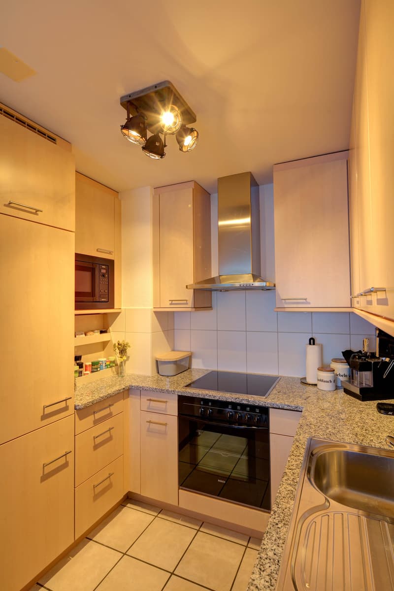 Modern kitchen with granite countertop, stainless steel sink, built-in oven, under-cabinet lighting, wooden cabinets, microwave, and open shelving with kitchenware.