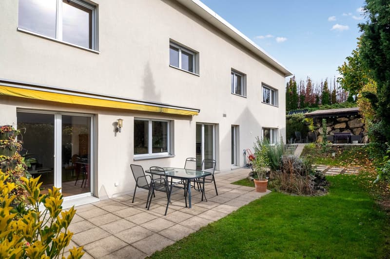 Beige house, large windows, sliding doors, paved patio with table and chairs, awning, garden with plants