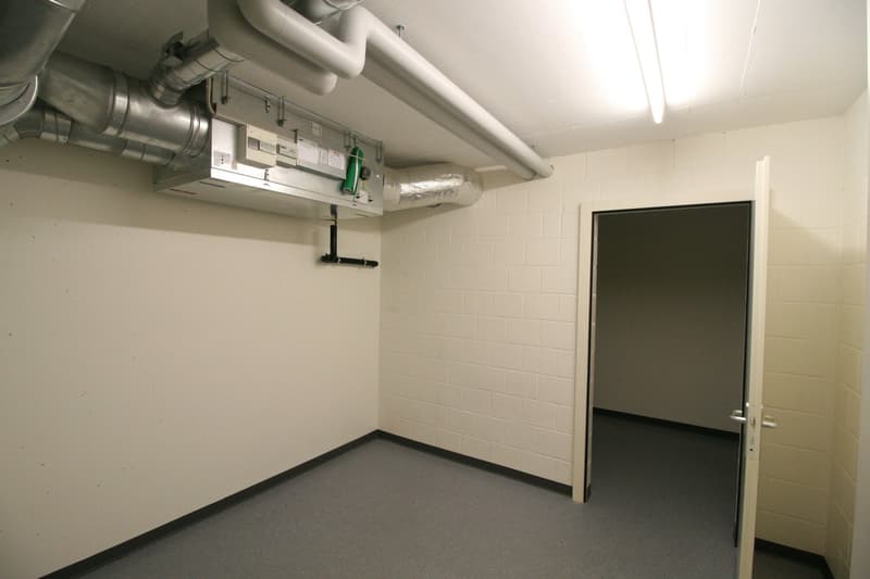A utility room with pipes and ducts on the ceiling, an open door, brick walls, and a gray floor.