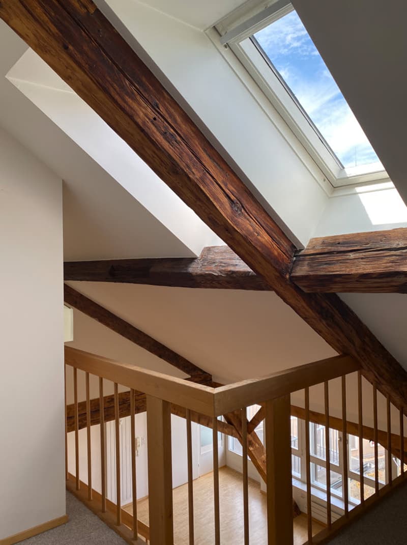 The image shows a loft or attic space with exposed wooden beams and a skylight window. The space features a wooden railing and an open layout.