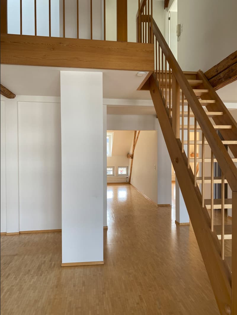 The image shows the interior of a house or apartment. It features a wooden staircase with a railing, wooden beams on the ceiling, and a hardwood floor. The space appears to be open-concept, with a hallway leading to other rooms. The walls are painted whit