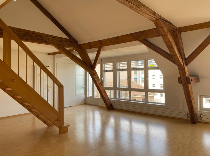 Spacious open-plan living area with exposed wooden beams, large windows providing natural light, and a balcony or terrace visible through the windows.