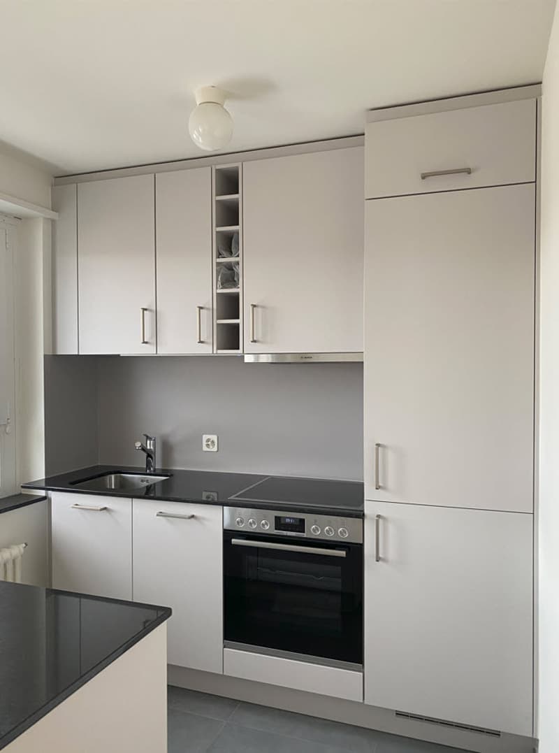 The image shows a modern, minimalist kitchen with white cabinets and drawers. The kitchen features built-in appliances, including an oven and a dishwasher, which is visible in the image. The countertops are made of a dark material, likely granite or quart