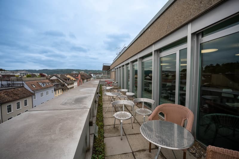 Outdoor seating area with tables and chairs, concrete floor, metal railing, glass walls, city view