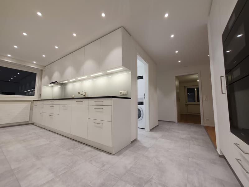 Modern, open-concept kitchen with white cabinets, black countertops, built-in appliances including a dishwasher and washing machine, and recessed lighting throughout the space.