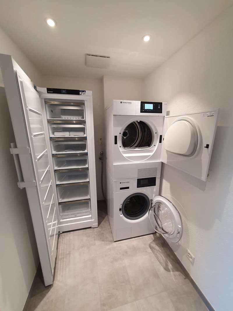 The image shows a utility room with a refrigerator, a washing machine, and a dryer. The room has white walls and a tiled floor.