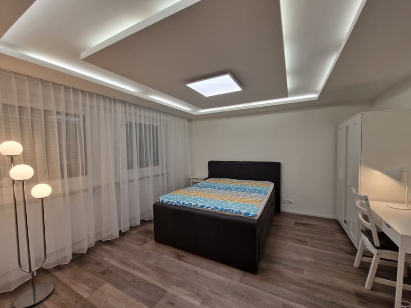 Spacious bedroom with hardwood flooring, white walls, large windows with sheer curtains, a black upholstered bed, and a small table with chairs near the window.