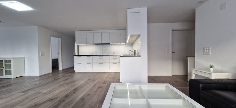 Spacious open-plan living area with modern white kitchen, hardwood flooring, and glass coffee table